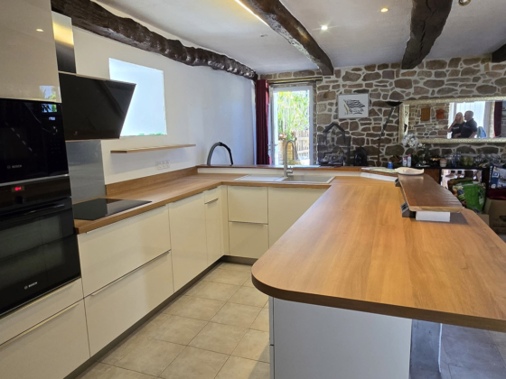 Vue d'ensemble d'une cuisine contemporaine blanc et bois sous un plafond poutres apparentes à Hennebont - Décobois
